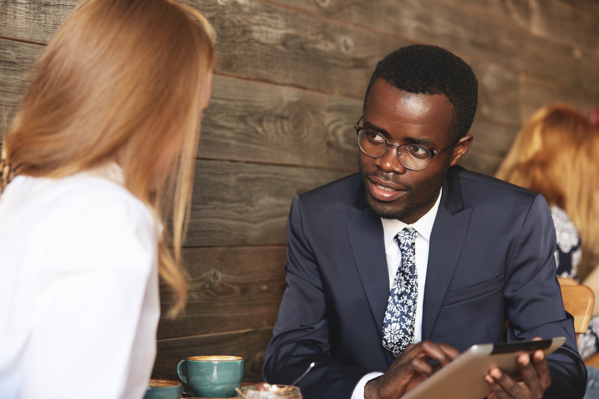 Imagem de homem negro de óculos e terno, segurando um tablet e conversando com mulher loira sobre como um software pode favorecer uma procuradoria jurídica