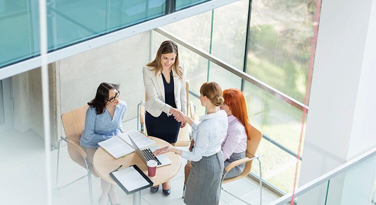 Imagem de mulheres reunidas, duas de pé apertando as mãos, e duas sentadas para ilustrar dicas para melhorar o networking do escritório de advocacia
