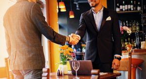 Imagem de dois homens de pé, vestindo terno, apertando as mãos em um restaurante ilustrando um encontro com advogado correspondente