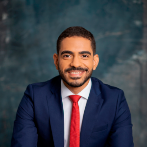 Foto de Bruno Paiva, homem pardo de barba e cabelo escuros vestindo um paletó