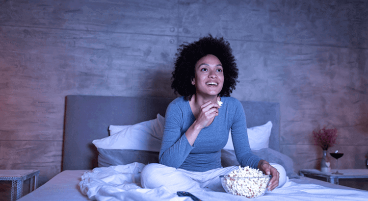 Mulher negra sentada na cama com um balde de pipoca em frente à TV assistindo séries de advogados